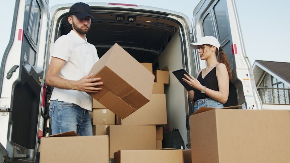 pexels-photo-5025667-5025667 Two delivery workers handling cardboard boxes by a van, checking orders on tablet.