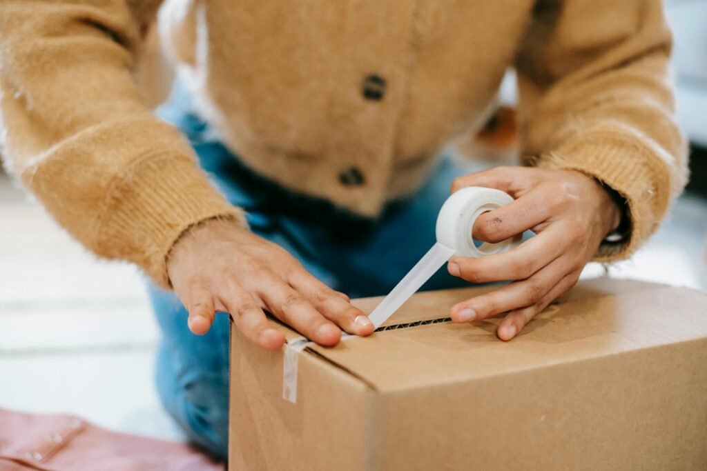 pexels-photo-6348095-6348095 A woman uses tape to seal a cardboard box at home, preparing for shipping or moving.